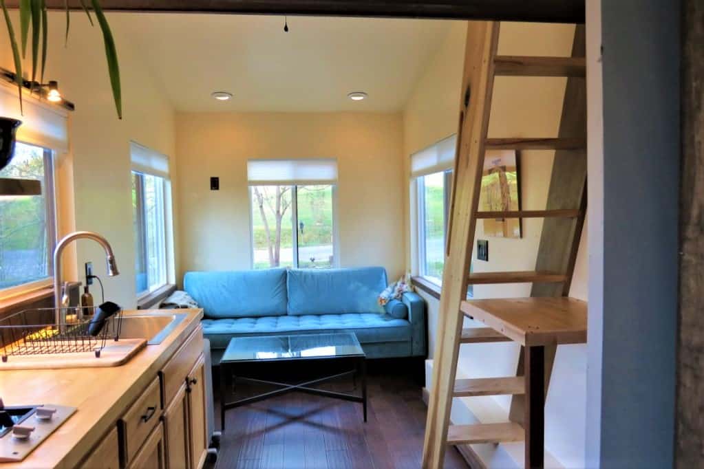 Interior of tiny home with small kitchen, blue couch and ladder to loft with bed.