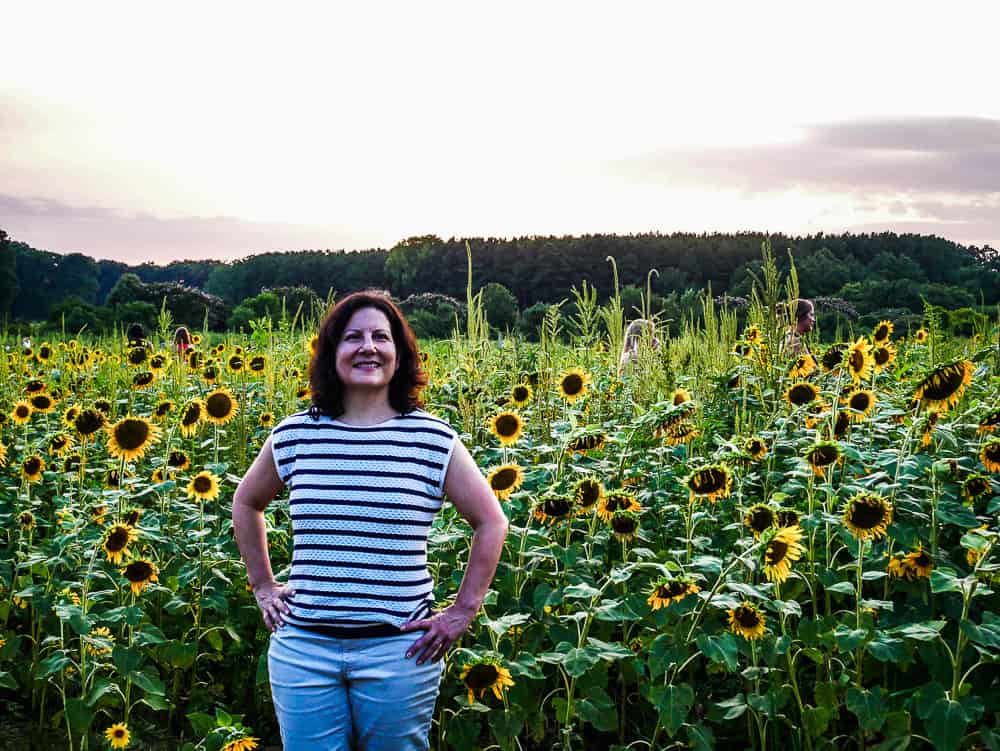 Author standing in front of sunflowers in white and blue striped shirt.