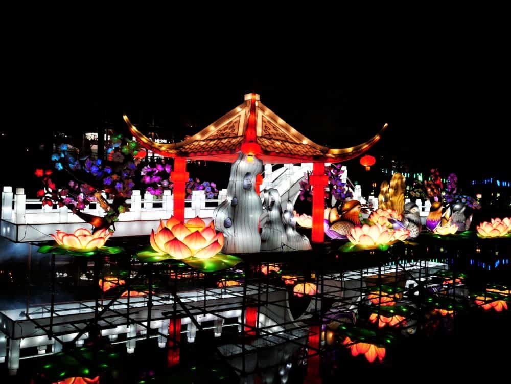 A lantern scene of a park with a pagoda and white bridge surrounded by flowers on a lake.