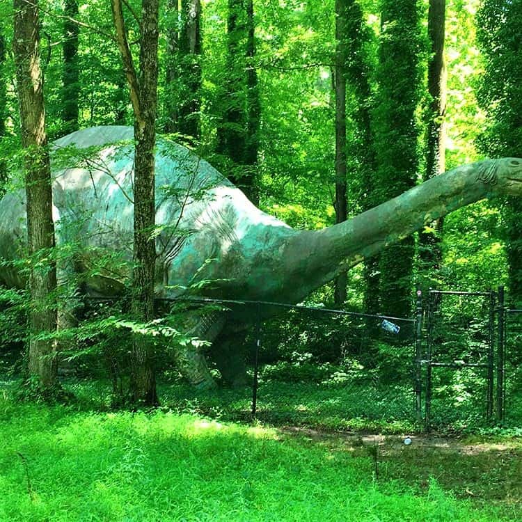 Large green dinosaur statue in a wooded are between trees with a small metal gate in front of it. 