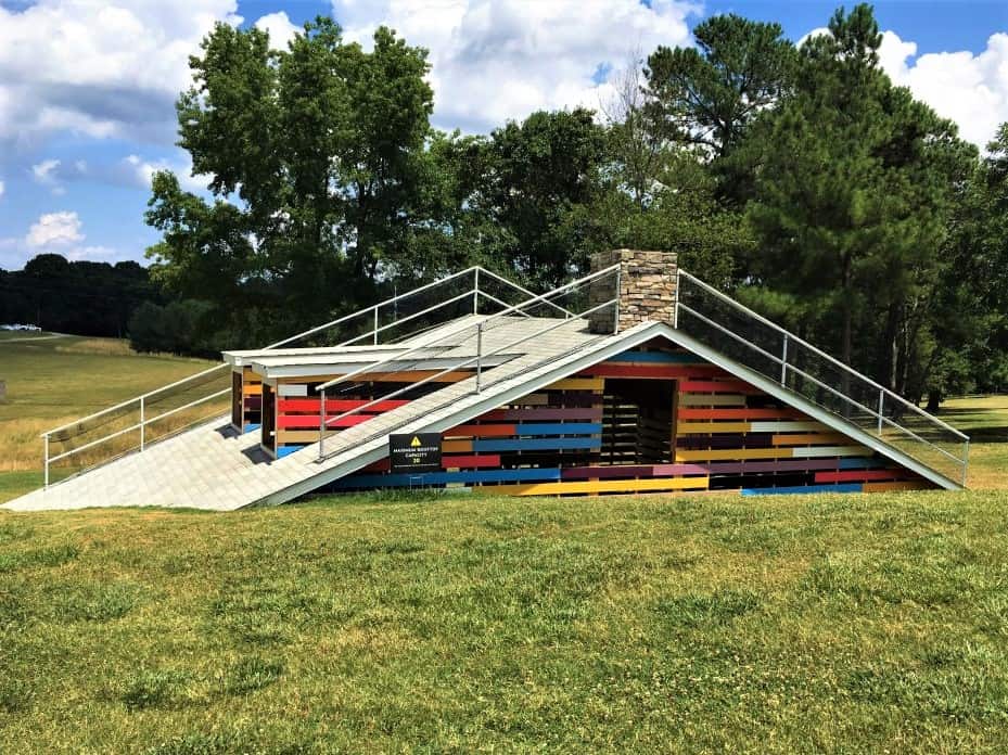 What looks like the roof of a house coming out of the ground. It is an a-frame roof with bright colored slats and a small chimney. 