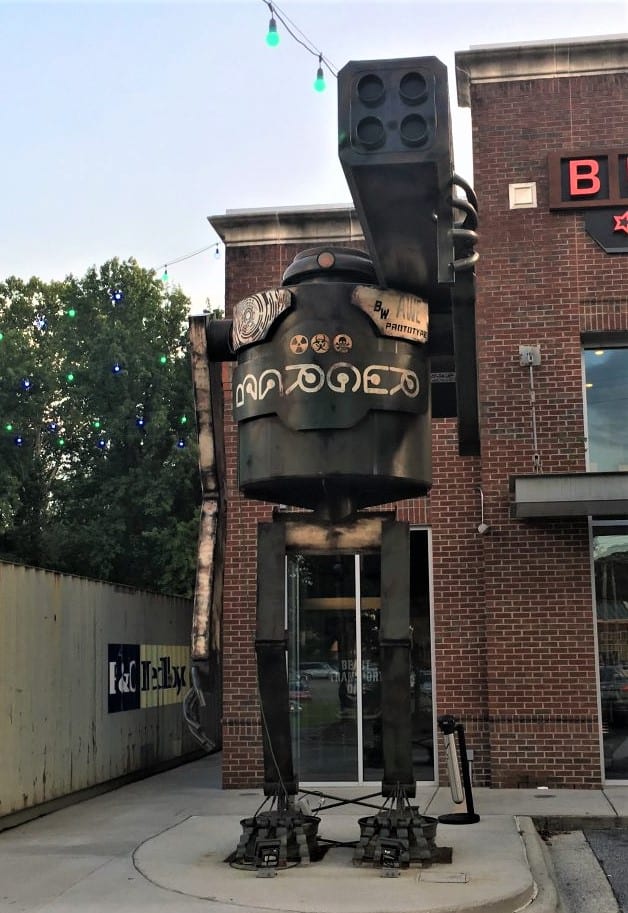 Metal robot that is over 20 feet tall. It has tall legs and a round, barrel top. The robot is holding a large metal gun. It is in front of a 2-story brick building. 
