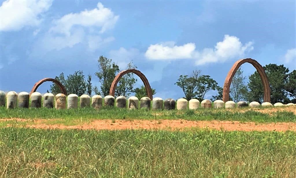 Three large copper colored rings getting larger from left to right with small stone pillars in front of them.  There is a blue sky with clouds in the background and green grass in the front. 