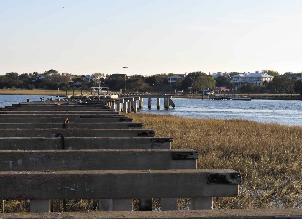 The remaining pilings and frame of the old Pitt Street Bridge. There is nothing left but wooden pilins with a wooden bar across them leading into the water. The bridge does not make it to the other side. 