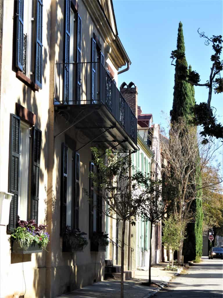 A view of houses on a street in Charleston. They are two story Victorian looking and attached. The first is a muted yellow, followed by a house with green shutters, then a pink house.  
