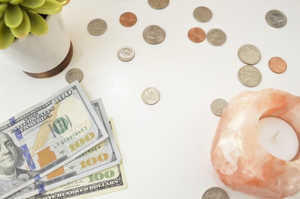 White surface with $100 dollar bills on the left, a white candle in a pink holder, green plant in a white vase, and coins scattered around.