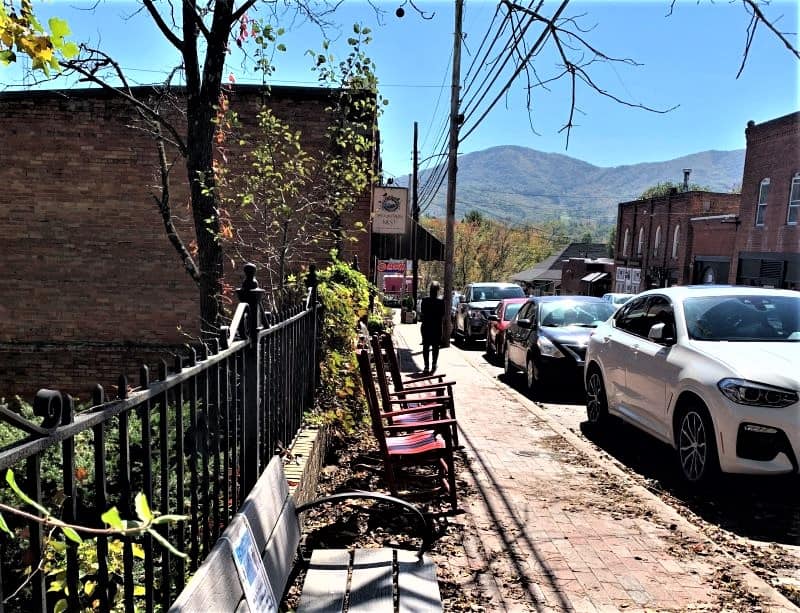 A sidewalk in downtown Black Mountain with a small bench and three rocking chairs by a black iron fence. A woman is walking past the chairs to a store further down the block. There are mountains in the background. 