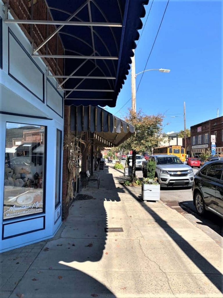 View of street from the sidewalk The stores all have black awnings and there are cars parked along the street. The first building is a bright blue. 