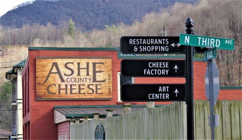 Street sign indicating Restaurants and Shopping, Cheese Factory, and Art Center on Third Ave in West Jefferson. Behind the sign is the red brick side of the Ashe County Cheese store with their sign viewable. 