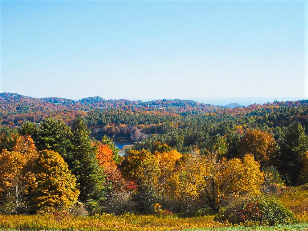 Colorful fall trees surrounding the Bass Pond at Moses Cone Memorial Park with mountains of colorful trees in the background.