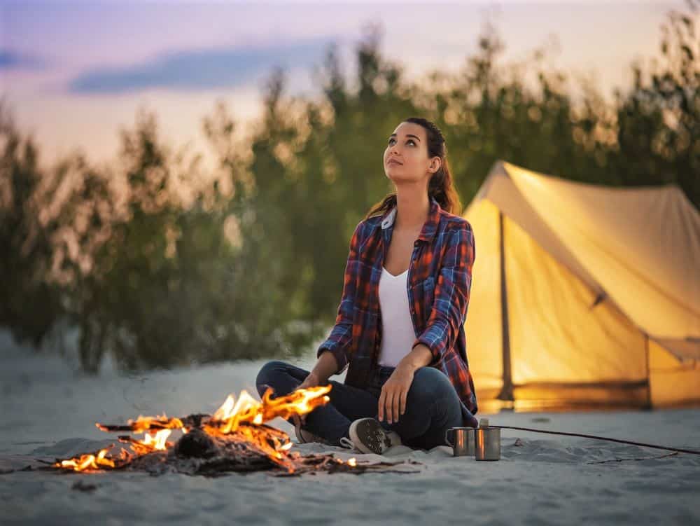 Woman camping with yellow tent behind her. She is sitting in front of a fire.