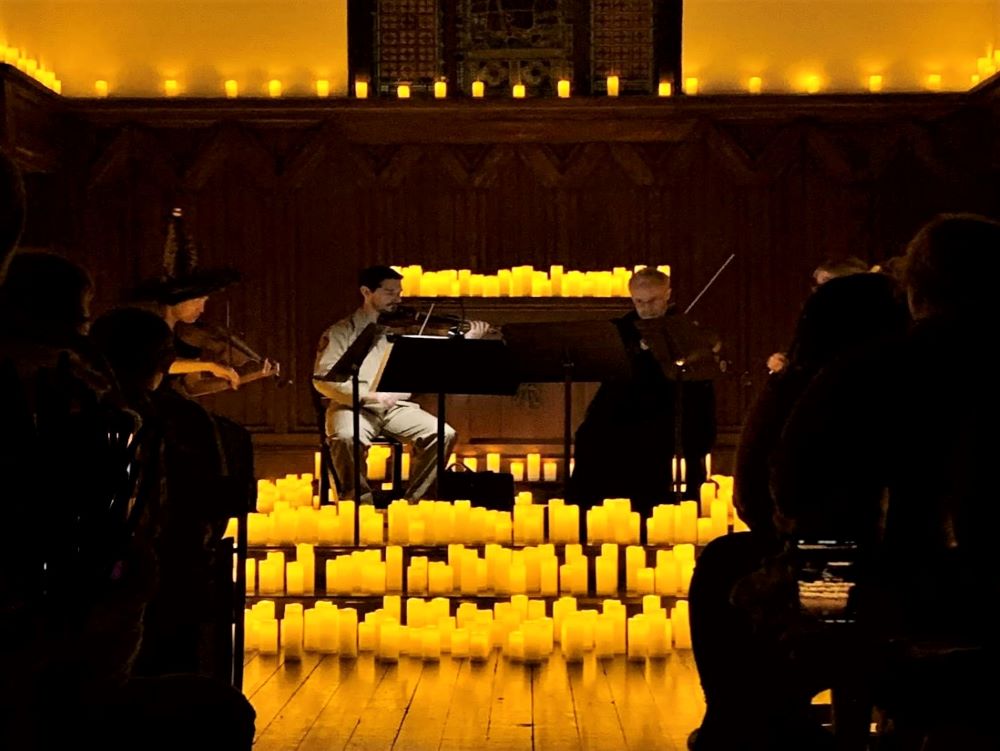 A small string quartet on a stage with battery operated candles in three rows in front of them and other candles behind them and on a ledge in the background. 