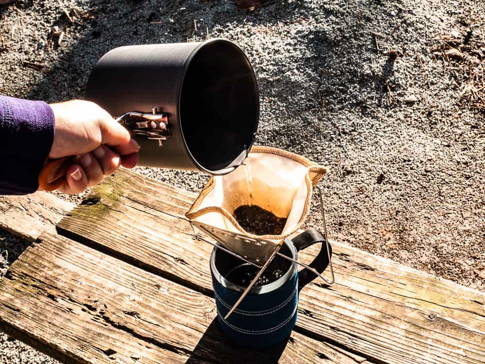 Metal collapsible pour over with coffeesock inside full of wet coffee grounds sitting on top of blue travel mug with water being poured over the coffee.