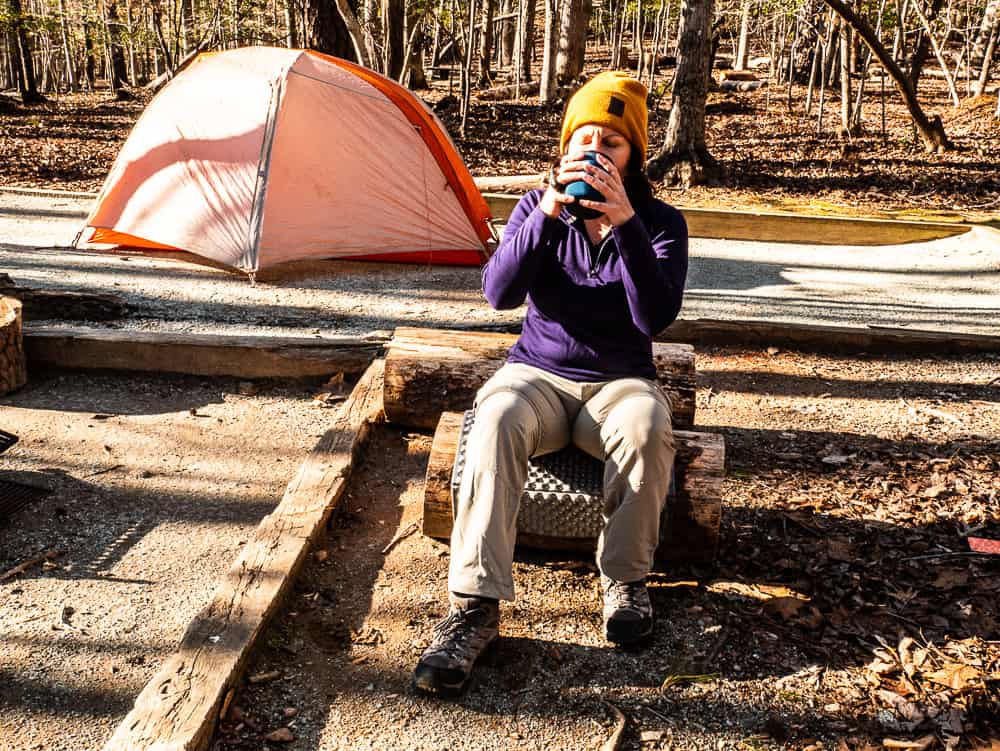 Woman sitting on log drinking coffee from a blue mug with tent in the background.