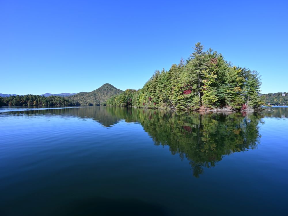 Dark blue lake with small island of trees in the middle and mountains in the background. 