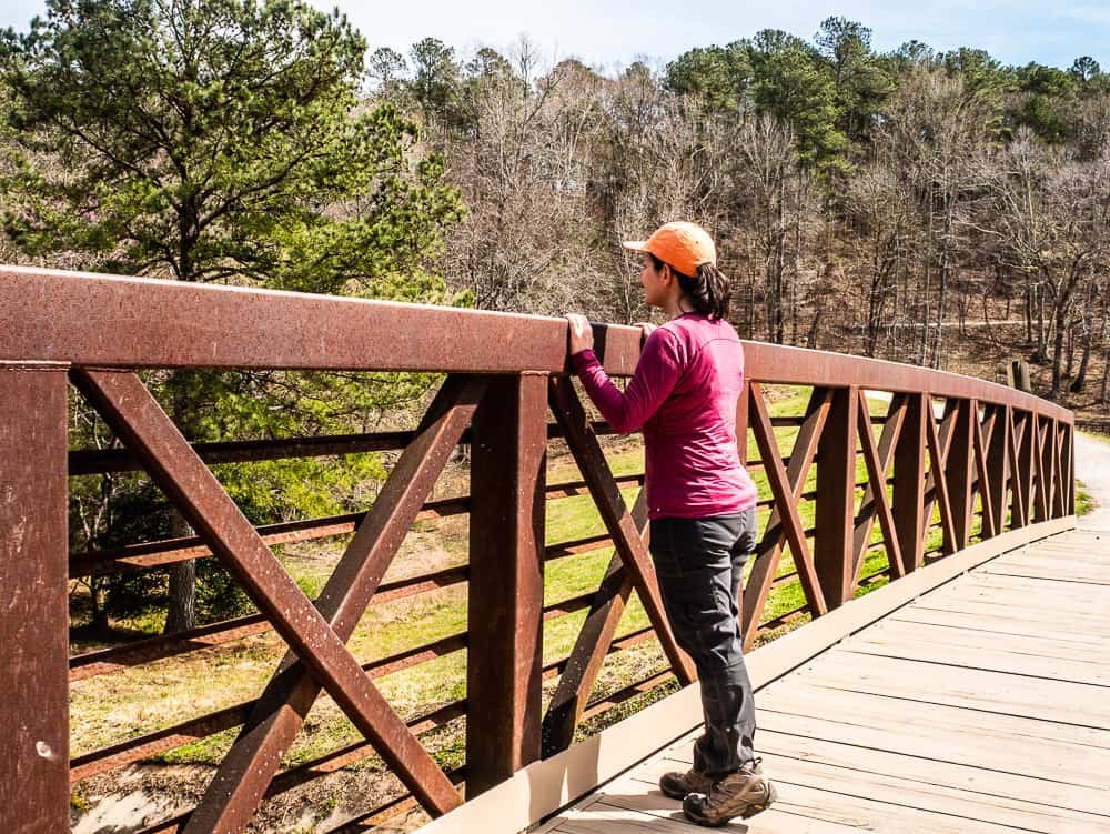 Woman in red shirt and orange hat standing on side of small bridge, looking over the side. 