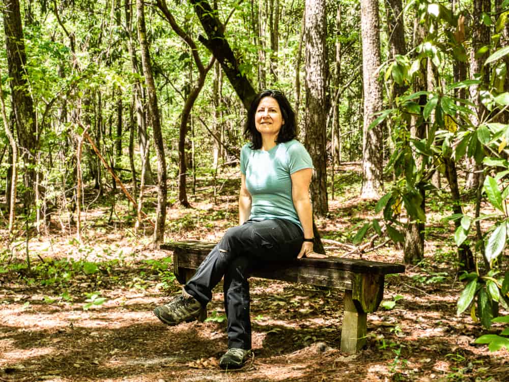 Woman in blue shirt and black pants sitting with leg crossed on a wooden bench surrounded by pine trees.
