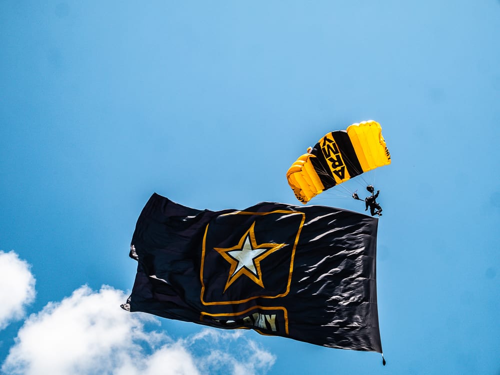 Skydiver with yellow and black parachute that says army. He is trailing a large black flag with a star that says US Army. 