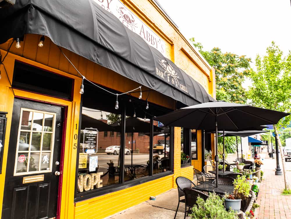 Yellow building with black trim and black awning with metal table and chairs topped with black umbrellas for eating.