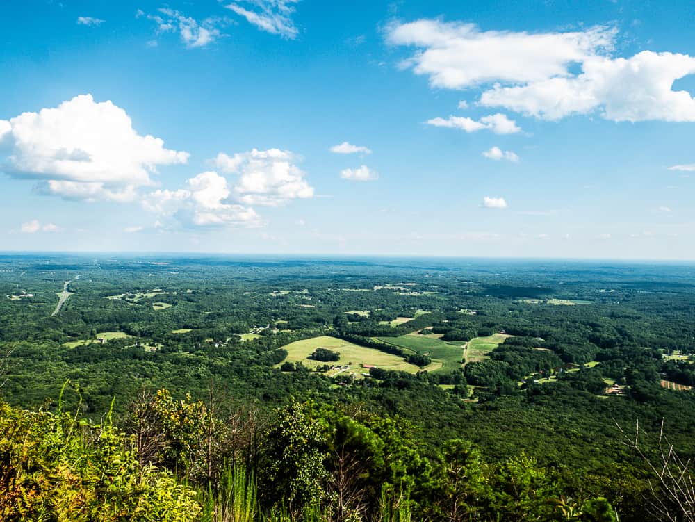 View of surrounding farm land from top of Pilot Mountain. Green trees and areas that have been cleared with houses with a blue sky.