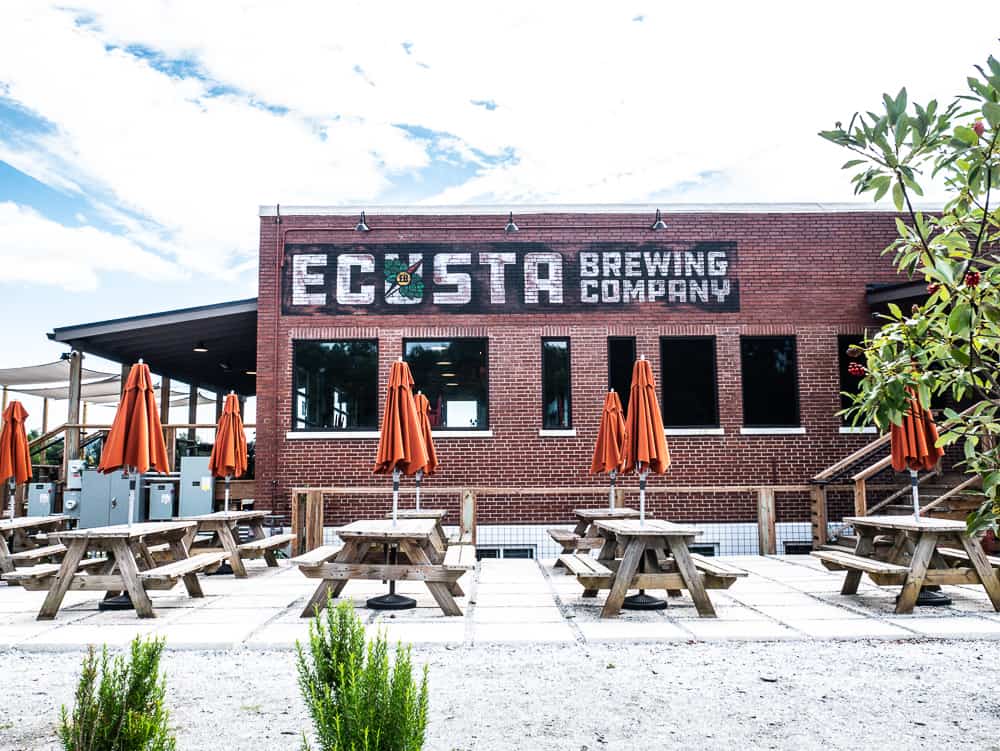Old red brick warehouse with picnic tables in front and sign for Ecusta Brewing Company painted in white on the side. 