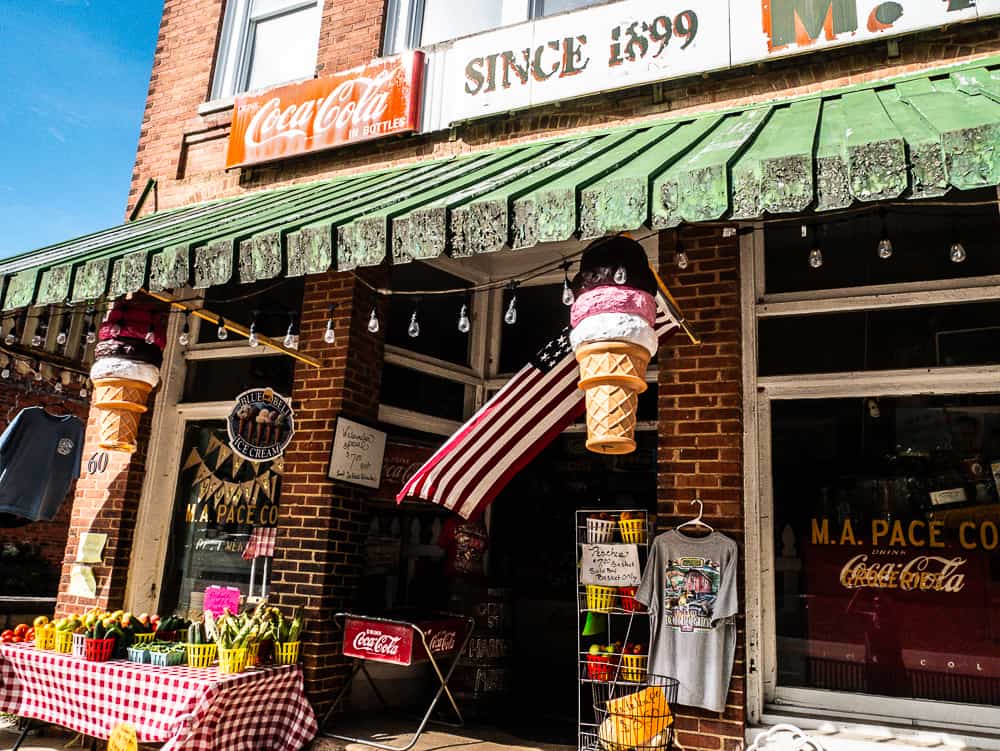Red brick store with green awning and coca cola sign with Since 1899. In the front are baskets of apples and a large, plastic ice cream cone with an American Flag behind it. 
