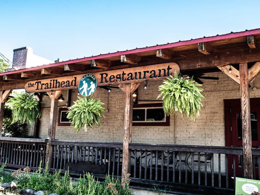 One story beige brick building with ferns hanging from the awning and a sign for The Trailhead Restaurant. 