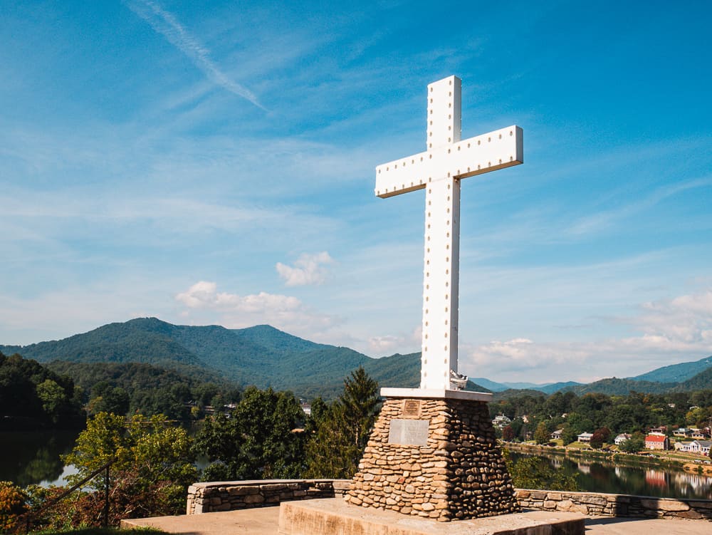 White cross with lights around the edges sitting on a rock pedestal with a lake and mountains in the background.