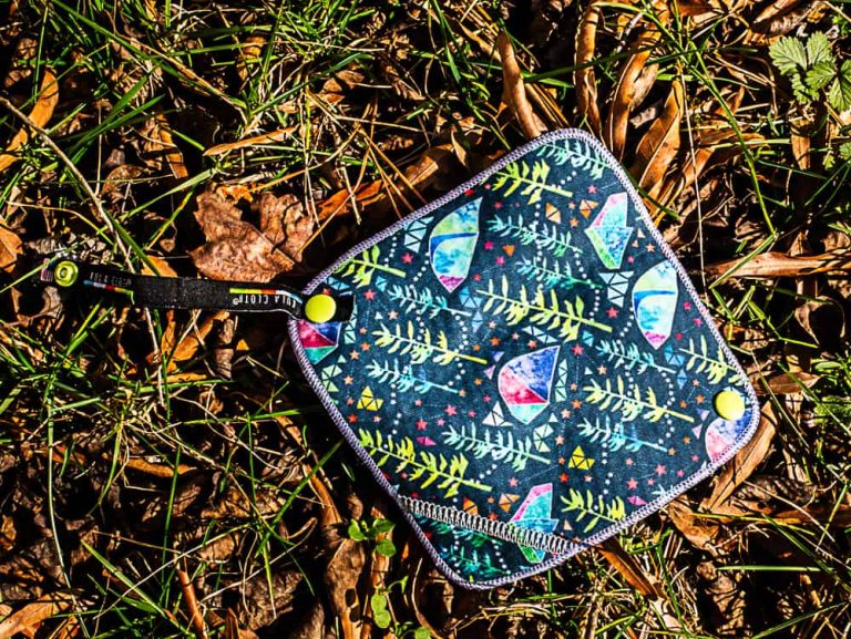 Square piece of cloth with green and blue trees and red and blue tents with a small strap that has a snap. It is sitting on grass and fallen leaves.