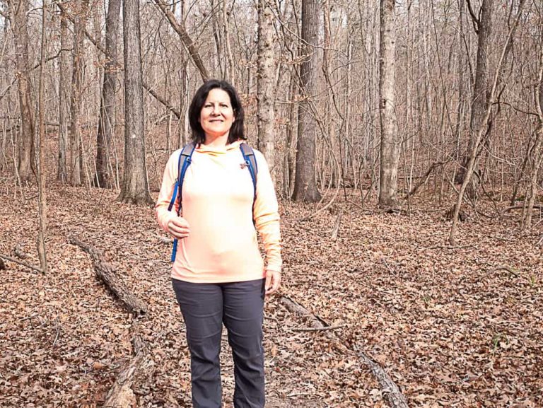Woman wearing peach long-sleeve sun shirt and grey pants with a blue day pack walking through woods with red and brown leaves on the ground.