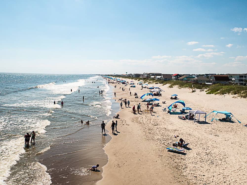 View of beach from above. A line of houses on the left are separated from the beach by reed-covered sand dunes. There are people on the beach in the sand, some under blue umbrellas. Other people are standing in the waves.
