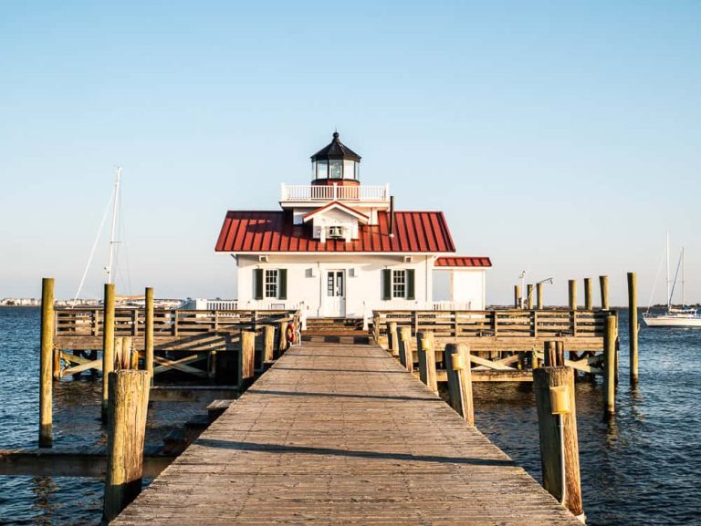 White building with red roof and small lighthouse on top. This is a screw-pile lighthouse called the Roanoke Marshes Lighthouse. There is a wooden pier that leads to the building which is floating in the water.
