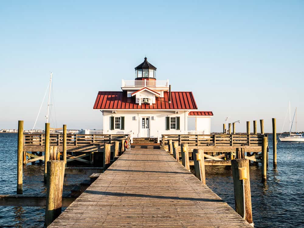 White building with red roof and small lighthouse on top. This is a screw-pile lighthouse called the Roanoke Marshes Lighthouse. There is a wooden pier that leads to the building which is floating in the water.