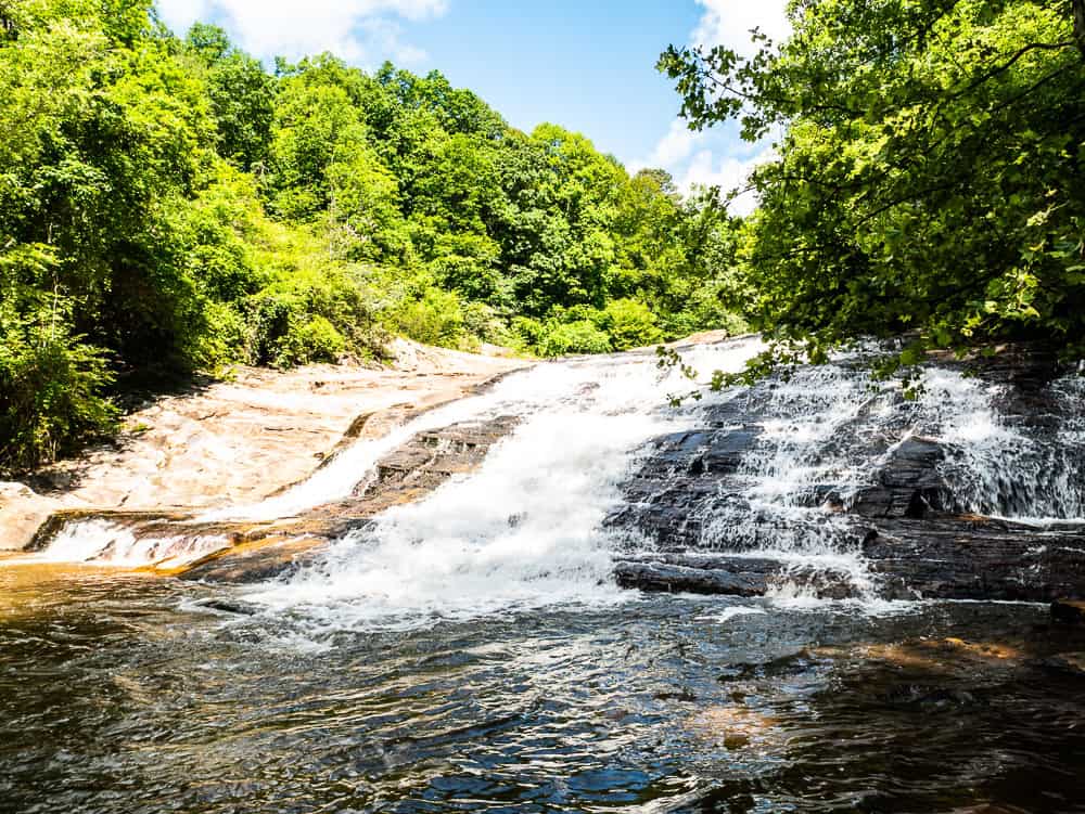 Short but wide waterfall cascading down brown rocks into a pool at the bottom. Both sides are lined with lush green trees. 