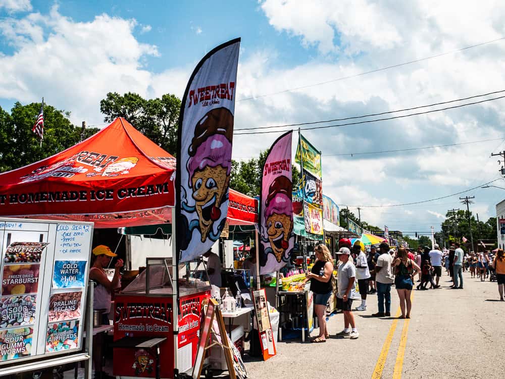 Several colorful tents along a street with vendors selling food. People are lined up in front of the tents. The front one is a red tent with letters showing is sells homemade ice cream. There are large flags in front with an ice cream cone cartoon character. 