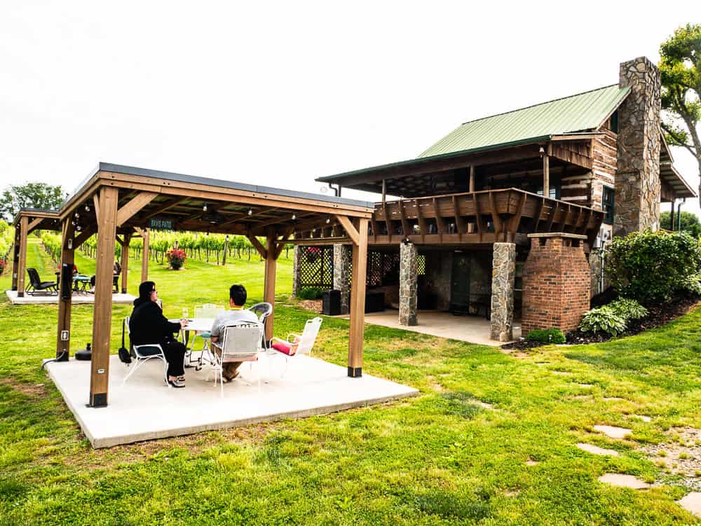 Two people sitting at a table under a covered gazebo with a two story log cabin to their right. 