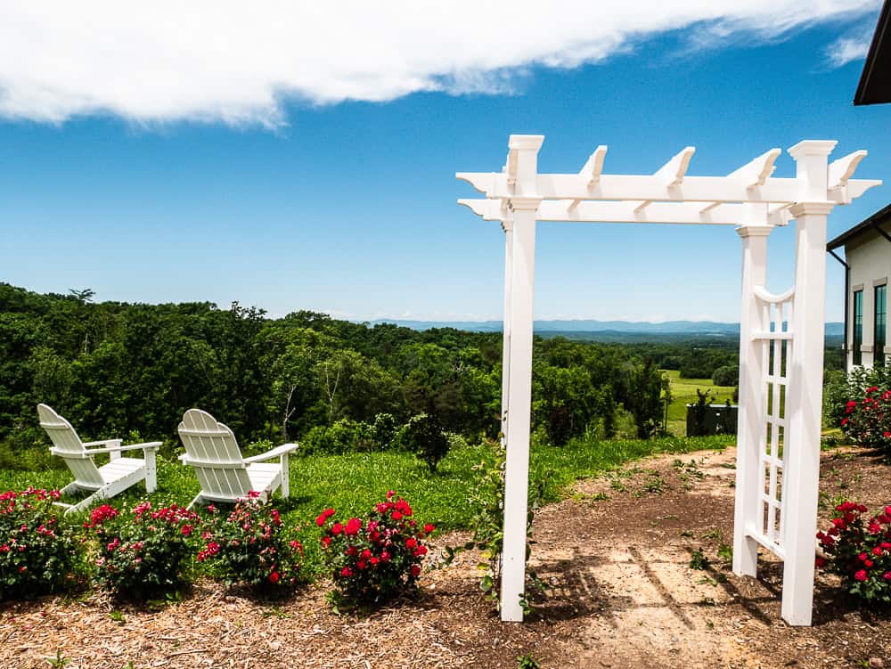 A white gazebo entrance with two white Adirondack chairs overlooking vineyard fields and the mountains.
