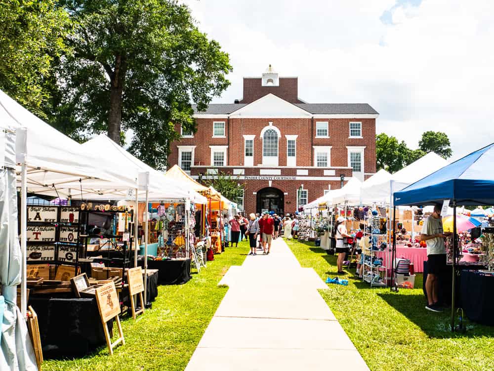 Vendors under white tents with one blue tent. They are pitched in the grass on either side of a sidewalk leading to a red brick colonial building that serves as the County Courthouse. 