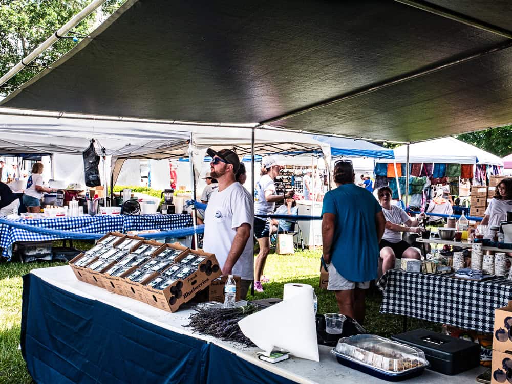 Man standing under a tent selling packs of blueberries. There are several pint containers in wooden boxes. Other vendors under tents are in the background. 