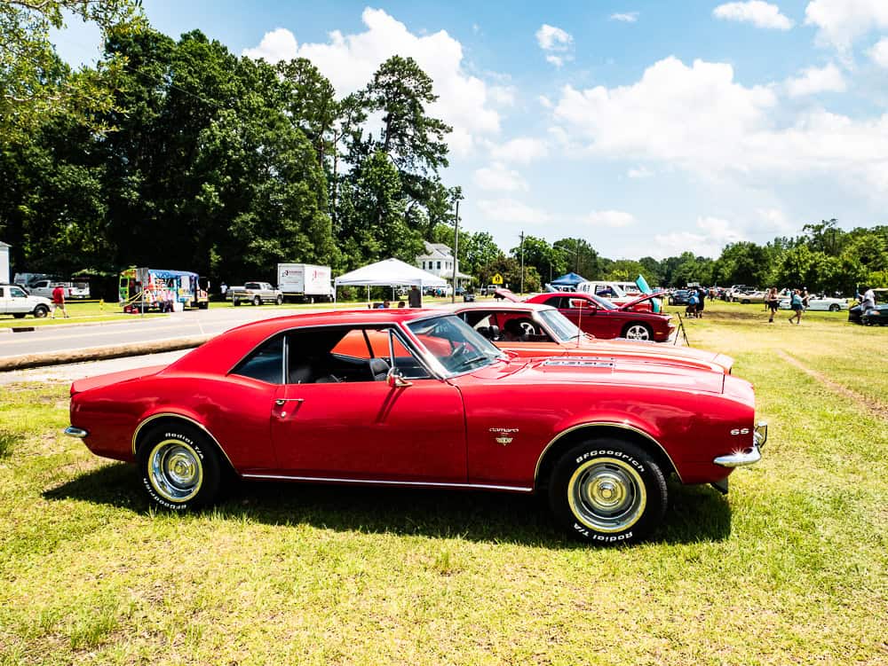 Red classic car with wheels that have yellow along the inside rim. There are other red cars behind it. They are parked in a grassy field. 