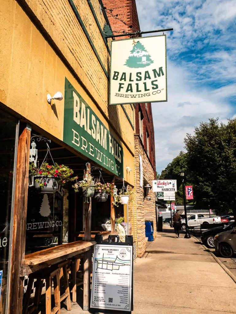 Vertical photo of brown-yellow storefront with green sign for Balsam Falls Brewing. There is a small eating are in front and a large menu on the sidewalk. 