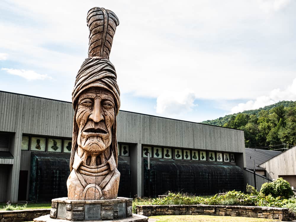 Large wooden carving of a Cherokee Indian that stands over 25 feet tall. Behind him is a gray building that houses the Museum of the Cherokee Indians.