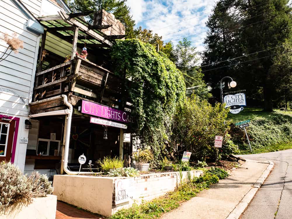 White wooden building with a wooden porch on the second floor. Green leaves cascade down from the porch to the first floor. A pink banner with white text says City Lights Cafe. 