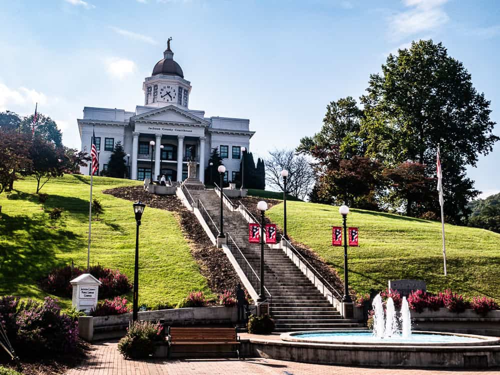 White two story building up a steep hill with a staircase leading to it. There is a fountain at the bottom of the staircase. The building looks similar to the Whitehouse and has a clock tower on the top.