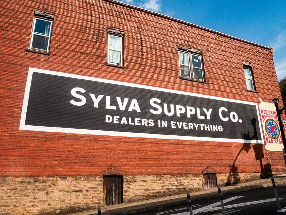 Two story red brick building with windows on the second floor. The first floor is covered by a mural for Sylva Supply Co. Dealers in Everything. 
